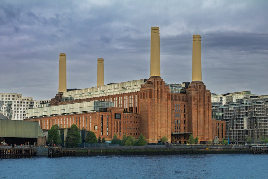 The image displays the iconic Battersea Power Station, a large historic industrial building featuring a red brick facade and four tall beige chimneys extending upwards against a cloudy sky. The structure's exterior showcases a combination of brickwork and extensive glass windows, with some sections appearing modernized with glass extensions along the sides. The power station is situated by a waterfront, with a visible water body in the foreground reflecting parts of the building and the cloudy sky above. To the left, there is a small wooden pier or jetty with a few people standing on it, adjacent to a green-roofed warehouse or boathouse. Surrounding the power station are modern residential and commercial buildings with glass and steel facades, contrasting the historic industrial architecture. The scene appears to be taken during daylight, with diffuse natural light illuminating the scene evenly. The image, associated with rubbish removal services such as those provided by Rubbish Removal Battersea, subtly highlights the blend of old industrial sites and the modern urban environment, where alternative waste handling and site clearance might be relevant in proximity to such landmarks.