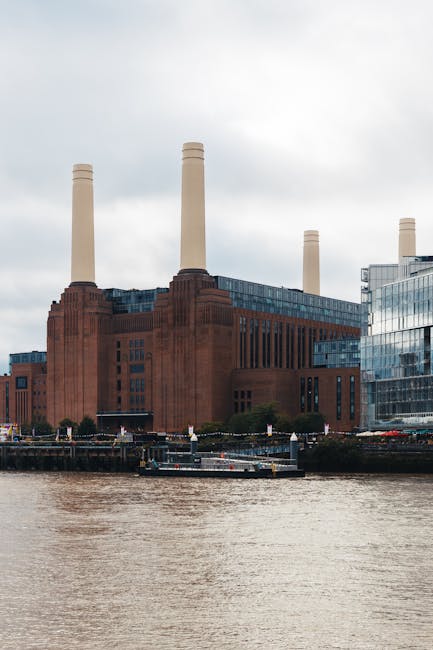 A large industrial building with a prominent red brick facade and narrow vertical windows, situated along the riverbank. The structure features four tall, cylindrical smokestacks in a beige color extending above the roofline. To the right of the building, there are modern glass-fronted offices or commercial spaces reflecting the cloudy sky. In the foreground, the river's water appears brownish and slightly rippled, with a small boat docked along the wharf area. The environment suggests an urban riverside setting with a mix of historic industrial architecture and contemporary development. This scene relates to private waste handling or on-site clearance, as it shows a prominent power station or factory-like building that could be associated with waste management or industrial site clearance. Light overcast lighting creates a neutral, subdued atmosphere, emphasizing the structural details and materials of the building without harsh shadows. Such locations often require specialised rubbish removal services, such as those offered by Rubbish Removal Battersea, to manage waste from redevelopment or maintenance activities related to utility or industrial facilities.
