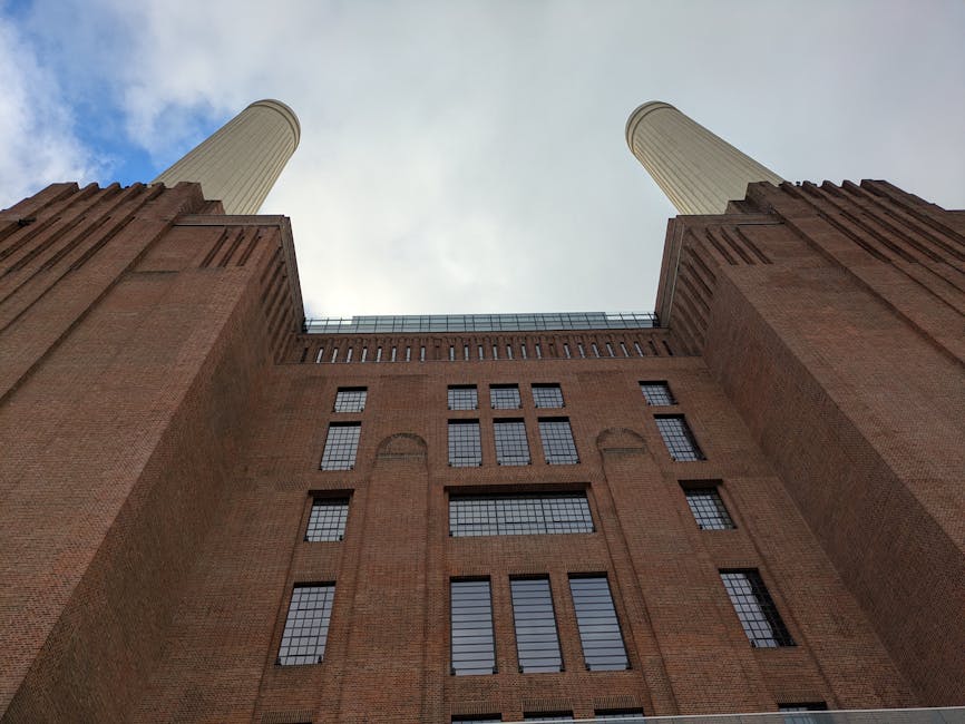 The image displays the iconic Battersea Power Station, a large historic industrial building featuring a red brick facade and four tall beige chimneys extending upwards against a cloudy sky. The structure's exterior showcases a combination of brickwork and extensive glass windows, with some sections appearing modernized with glass extensions along the sides. The power station is situated by a waterfront, with a visible water body in the foreground reflecting parts of the building and the cloudy sky above. To the left, there is a small wooden pier or jetty with a few people standing on it, adjacent to a green-roofed warehouse or boathouse. Surrounding the power station are modern residential and commercial buildings with glass and steel facades, contrasting the historic industrial architecture. The scene appears to be taken during daylight, with diffuse natural light illuminating the scene evenly. The image, associated with rubbish removal services such as those provided by Rubbish Removal Battersea, subtly highlights the blend of old industrial sites and the modern urban environment, where alternative waste handling and site clearance might be relevant in proximity to such landmarks.