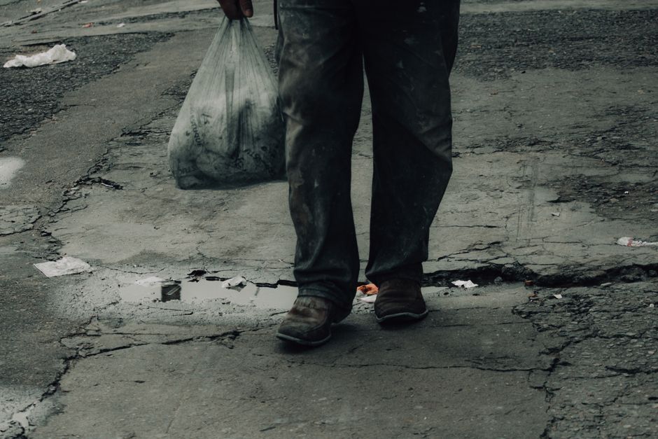 The image depicts the lower half of a person wearing dark, slightly dirty work trousers and sturdy brown work shoes, walking on a cracked asphalt surface. The individual is holding a large, semi-transparent plastic rubbish bag filled with waste, which appears to contain dark and light debris. The surrounding environment includes an uneven, weathered pavement with visible cracks and patches, as well as small scattered pieces of trash and paper remnants. In the background, the asphalt continues, with some fading lighting indicating an outdoor setting. The scene suggests a manual rubbish collection or clearance activity, associated with private waste handling services such as those offered by Rubbish Removal Battersea, emphasizing the physical aspect of waste disposal in an urban context, fitting naturally with the theme of alternative waste removal or on-site clearance processes.