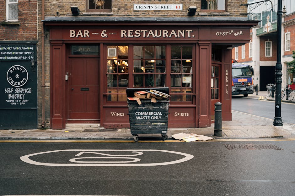 In front of a closed, dark red wooden storefront labeled as a bar and restaurant, a black waste disposal bin marked with 'Commercial Waste Only' stands on the pavement, overflowing with flattened cardboard boxes and miscellaneous waste. The bin is situated directly beneath a large window pane divided into multiple smaller sections, with warm interior lighting visible through the glass. To the left of the bin, a blackboard sign advertising a self-service buffet is mounted on the building's wall, while on the right, a black bollard protects the storefront from vehicle encroachment, with a tall street lamp nearby. Behind the bin, a grey truck is parked on the street, and further along the sidewalk, bicycles are secured to a stand. The scene captures an urban setting, with the pavement paved in grey concrete and the road marked with white paint, including a partially visible diesel symbol painted directly on the street surface. The overall environment appears to be a busy city area where private waste collection services like Rubbish Removal Battersea frequently manage rubbish removal for local businesses, especially for commercial waste disposal outside regular council services.