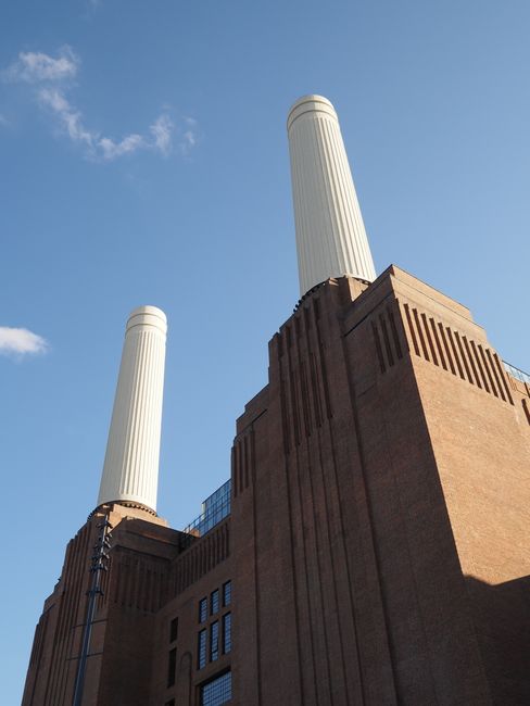A low-angle view of a large industrial building with two prominent white chimney stacks extending into a clear blue sky. The building is constructed from red-brown brickwork with vertical architectural details and rectangular windows visible on the facade. The chimneys are cylindrical with smooth surfaces and rounded tops, positioned side by side on the roof of the structure. The scene is illuminated by natural daylight, casting shadows on the building's surface, and the overall environment suggests an industrial area that may be involved in waste processing or energy generation. Such infrastructure typically signifies facilities where large quantities of waste or byproducts are managed, aligning with the context of alternative waste handling or private collection services like those offered by Rubbish Removal Battersea, situated in an urban setting with a focus on efficient, independent rubbish disposal.