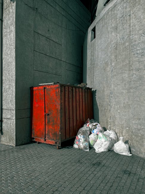 A large, weathered red metal skip with visible rust and peeling paint is positioned against a dark green wall on the left and a concrete wall on the right on a paved surface. The skip appears to be used for waste storage and is partially filled, with several transparent plastic bags of mixed waste, including packaging, paper, and household rubbish, placed directly on the ground beside it. The bags are loosely tied and vary in size, with some bulging and others more compact. The environment suggests an outdoor urban or commercial space, likely a back alley or service area, with minimal natural light highlighting the texture of the concrete and metal surfaces. This scene aligns with the type of privately arranged rubbish collection or waste disposal that companies like Rubbish Removal Battersea facilitate, providing an alternative to local council pickup for bulky or commercial rubbish. The setting emphasizes the importance of proper waste management and regulated disposal methods in urban environments.