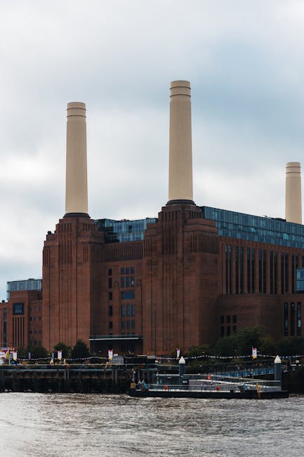 A large industrial building with a reddish-brown brick exterior and multiple vertical windows is situated along a riverbank. The structure features three prominent tall, beige smokestacks extending upward from the roof, with a textured surface and rounded tops. In the foreground, there is a body of water with a small, flat-bottomed barge or boat floating near the edge, which appears to be used for maintenance or transport. The riverbank includes a narrow walkway or quay with some small barriers and industrial fixtures. The sky above is cloudy, casting diffused light over the scene, creating a neutral atmosphere appropriate for a description related to waste management and alternative disposal methods associated with rubbish removal services, such as those provided by Rubbish Removal Battersea. Surrounding vegetation is sparse, with a few low shrubs or trees near the building base, emphasizing the urban and functional environment of the site.