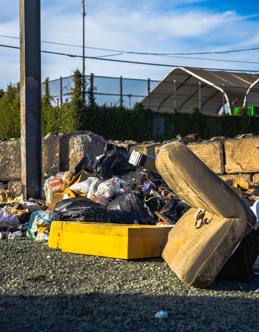 A large, weathered red metal skip with visible rust and peeling paint is positioned against a dark green wall on the left and a concrete wall on the right on a paved surface. The skip appears to be used for waste storage and is partially filled, with several transparent plastic bags of mixed waste, including packaging, paper, and household rubbish, placed directly on the ground beside it. The bags are loosely tied and vary in size, with some bulging and others more compact. The environment suggests an outdoor urban or commercial space, likely a back alley or service area, with minimal natural light highlighting the texture of the concrete and metal surfaces. This scene aligns with the type of privately arranged rubbish collection or waste disposal that companies like Rubbish Removal Battersea facilitate, providing an alternative to local council pickup for bulky or commercial rubbish. The setting emphasizes the importance of proper waste management and regulated disposal methods in urban environments.