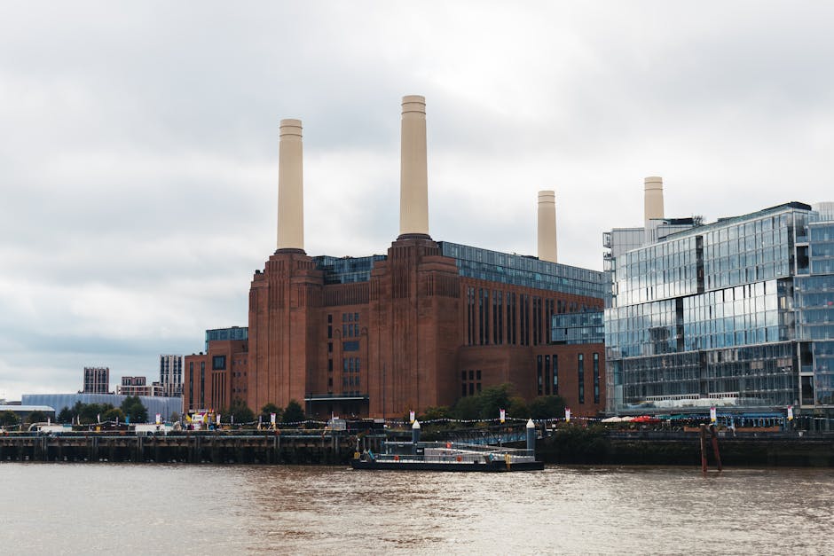 A large historic brick building with four tall, beige chimneys extending upward, situated along a riverbank. The building has a reddish-brown stone façade with vertical architectural details and multiple rows of windows on its upper levels. To the right of the historic structure, there is a modern glass building with reflective windows, creating a contrast with the brickwork. In the foreground, the river's brownish water is visible, along with a small docked boat or barge positioned near the shoreline. The sky overhead is cloudy with a diffuse light casting minimal shadows, emphasizing the industrial and urban environment and subtly suggesting the site’s potential relevance to private waste handling or on-site clearance activities associated with the cityscape. Rubbish removal services such as those offered by Rubbish Removal Battersea may operate in areas around such historic and modern structures to manage waste disposal in the region.