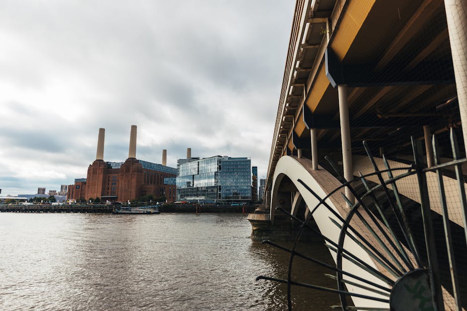 An urban waterfront scene showing the underside of a bridge or walkway on the right side, supported by vertical metal columns, with a section of black metal railing visible in the foreground. In the background, a large industrial building with red brick facades and four tall, cylindrical chimney stacks extends along the riverbank. Adjacent to the industrial structure is a modern glass office building with reflective windows. The river in the foreground has a slight ripple effect, and the overcast sky above is filled with gray clouds, creating a subdued lighting atmosphere. The scene reflects a typical cityscape with elements of industrial and modern architecture close to a waterway, which is relevant to the context of waste and rubbish removal, particularly in relation to urban infrastructure and the potential for private waste collection or disposal services in a city environment.