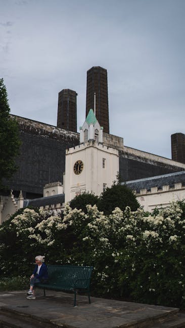A large historic brick building with four tall, beige chimneys extending upward, situated along a riverbank. The building has a reddish-brown stone façade with vertical architectural details and multiple rows of windows on its upper levels. To the right of the historic structure, there is a modern glass building with reflective windows, creating a contrast with the brickwork. In the foreground, the river's brownish water is visible, along with a small docked boat or barge positioned near the shoreline. The sky overhead is cloudy with a diffuse light casting minimal shadows, emphasizing the industrial and urban environment and subtly suggesting the site’s potential relevance to private waste handling or on-site clearance activities associated with the cityscape. Rubbish removal services such as those offered by Rubbish Removal Battersea may operate in areas around such historic and modern structures to manage waste disposal in the region.